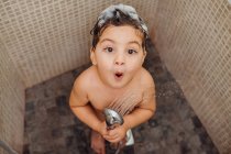 Desde arriba sonriente niño pequeño con espuma en la cabeza de pie en el baño con ducha y cantando mientras mira a la cámara - foto de stock