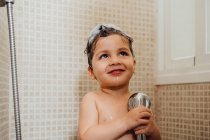 Bambino sorridente con schiuma sulla testa in piedi in bagno con doccia e canto mentre distoglie lo sguardo — Foto stock