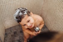 Зверху усміхнена маленька дитина з піною на голові стоїть у ванній кімнаті з душем і співом, дивлячись вниз — стокове фото
