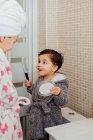 Adorable child in bathrobe standing in bright bathroom together with mother and combing wet hair after shower while looking in mirror — Stock Photo