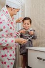 Вид збоку на матір в халаті та рушнику, що розчісує вологе волосся маленької дитини, стоячи у сучасній ванній кімнаті — стокове фото