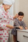 Seitenansicht der Mutter mit Bademantel und Handtuch Turban, die das nasse Haar eines kleinen Kindes kämmt, während sie im modernen Badezimmer steht — Stockfoto