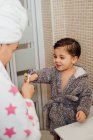 Симпатичный маленький мальчик в халате и улыбающаяся мать в полотенце тюрбан стоя в ванной комнате и чистя зубы — стоковое фото