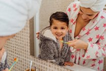 Petit garçon mignon en peignoir et mère souriante en serviette turban debout dans la salle de bain et brossant les dents — Photo de stock