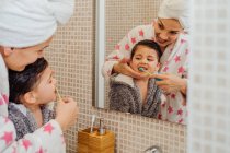 Симпатичний маленький хлопчик в халаті і усміхнена мати в рушнику турбан, що стоїть у ванній кімнаті і чистить зуби — стокове фото