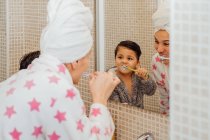 Симпатичный маленький мальчик в халате и улыбающаяся мать в полотенце тюрбан стоя в ванной комнате и чистя зубы — стоковое фото