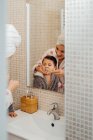 Cute little boy in bathrobe and smiling mother in towel turban standing in bathroom and brushing teeth — Stock Photo