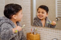 Criança adorável vestindo roupão aconchegante em pé no banheiro com escova de dentes e olhando no espelho — Fotografia de Stock
