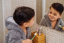 Adorable child wearing cozy bathrobe standing in bathroom with toothbrush and looking in mirror — Stock Photo