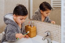 Очаровательный ребенок в удобном халате, стоящий в ванной комнате с зубной щеткой — стоковое фото