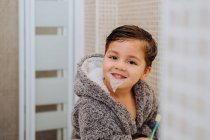 Criança adorável vestindo roupão aconchegante em pé no banheiro com escova de dentes — Fotografia de Stock
