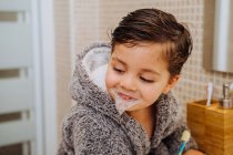 Adorable niño usando acogedor albornoz de pie en el baño con cepillo de dientes - foto de stock