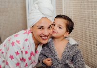 Веселая женщина с полотенцем тюрбан обнимает маленького ребенка в халате после принятия душа и глядя в камеру — стоковое фото