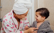 Mujer alegre con turbante de toalla abrazando a un niño pequeño en albornoz después de tomar una ducha y mirarse entre sí - foto de stock