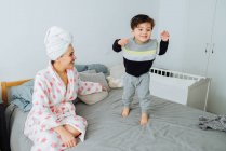 Mujer alegre en albornoz vestirse hijo pequeño mientras juegan juntos en casa mirándose el uno al otro - foto de stock