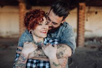 Positives junges Hipster-Paar mit Tätowierungen genießt die gemeinsame Zeit und umarmt sich, während es bei sonnigem Wetter vor schäbigen Steinbauten steht — Stockfoto