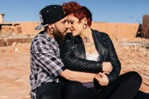 Positivo jovem casal hipster com tatuagens desfrutando de tempo juntos e abraçando enquanto de pé contra a construção de pedra gasto em dia ensolarado — Fotografia de Stock
