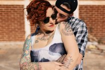 Позитивна молода пара хіпстерів з татуюваннями насолоджується часом разом і обіймається, стоячи проти кам'яної конструкції в сонячний день — стокове фото