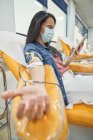 Низкий угол обзора молодой женщины в защитной маске, просматривающей смартфон во время процедуры переливания крови в больнице — стоковое фото