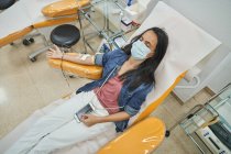 De cima de fêmea jovem relaxado em máscara protetora com smartphone sentado em cadeira médica durante doação de sangue no hospital moderno — Fotografia de Stock