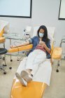 Vista lateral de ángulo bajo de la mujer joven en el teléfono inteligente de navegación máscara protectora durante el procedimiento de transfusión de sangre en el hospital - foto de stock