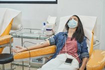 Von oben eine entspannte junge Frau in Schutzmaske mit Smartphone, die während der Blutspende im modernen Krankenhaus im Behandlungsstuhl sitzt — Stockfoto