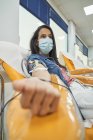 Вид сбоку молодой женщины в защитной маске, сидящей в медицинском кресле во время процедуры переливания крови в современной больнице — стоковое фото
