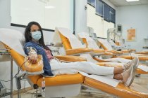 Vista lateral de la joven en máscara protectora sentada en sillón médico durante el procedimiento de transfusión de sangre en el hospital contemporáneo - foto de stock