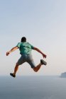 Vista posteriore ad angolo basso di un maschio irriconoscibile in abiti casual che salta in alto contro il cielo blu e il mare durante la sera d'estate — Foto stock