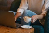 Cropped unrecognizable young female freelancer in casual clothes having tasty hot drink while sitting at round wooden table and using netbook in light modern cafe — Stock Photo