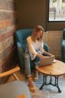 Junge blonde, glückliche Freiberuflerin in lässiger Kleidung mit leckerem Heißgetränk, während sie am runden Holztisch sitzt und Netbook in einem leichten modernen Café benutzt — Stockfoto