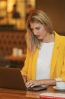Mulher de negócios adulta focada em desgaste formal olhando para longe enquanto sentada à mesa de madeira com laptop na loja de café contemporânea — Fotografia de Stock