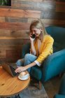 High angle of laughing adult female freelancer in casual clothes focusing on laptop screen and typing while sitting in soft blue armchair at round wooden table with cup of hot drink and making phone call in modern cafe — Stock Photo