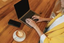 From above cropped unrecognizable young female freelancer in casual clothes having tasty hot drink while sitting at wooden table and using laptop in light modern cafe — Stock Photo
