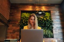 Знизу орієнтована доросла бізнес-леді в офіційному одязі, дивлячись далеко, сидячи за дерев'яним столом з ноутбуком в сучасному кафе — стокове фото