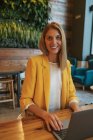 Erwachsene überglückliche Geschäftsfrau in formaler Kleidung blickt in die Kamera und lächelt, während sie an einem Holztisch mit Laptop in einem modernen Café-Shop sitzt — Stockfoto