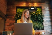 Erwachsene überglückliche Geschäftsfrau in formaler Kleidung schaut weg und lächelt, während sie an einem Holztisch mit Laptop in einem modernen Café-Shop sitzt — Stockfoto