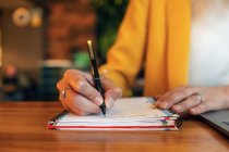 Обрізати невпізнавану бізнес-леді в елегантному барвистому одязі, пишучи з ручкою в блокноті, сидячи за дерев'яним столом і використовуючи ноутбук в сучасному офісі — стокове фото