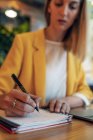 Empresária desfocada em roupas coloridas e elegantes que escrevem com caneta no caderno enquanto se senta na mesa de madeira e usa laptop no escritório contemporâneo leve — Fotografia de Stock