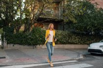Модная стройная блондинка-бизнесмен смотрит в элегантную ярко-желтую куртку и джинсы с цифровым планшетом, идущая одна по пешеходному переходу против экстерьеров жилых многоэтажных зданий и автомобилей, припаркованных на улице в центре города — стоковое фото