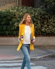 Trendy slim blond businesswoman looking away in elegant vivid yellow jacket and jeans with digital tablet walking alone along pedestrian crossing against exteriors of residential multistory buildings and cars parked on street in downtown — Stock Photo