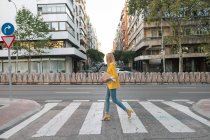 Side view of trendy slim blond female in elegant vivid yellow jacket and jeans with digital tablet walking alone along pedestrian crossing against exteriors of residential multistory buildings and cars parked on street in downtown — Stock Photo
