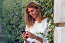 Mujer rubia relajada positiva con ropa casual enfocada en la pantalla y sonriendo mientras está de pie en la calle e interactuando con el teléfono inteligente cerca de la cerca cubierta de plantas trepadoras verdes - foto de stock