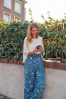 Positive relaxed blonde female in casual clothes focusing on screen and smiling while standing on street and interacting with smartphone near fence covered green climbing plant — Stock Photo