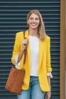 Jovem mulher loira feliz em casaco amarelo brilhante e jeans com bolsa marrom sobre o ombro sorrindo enquanto está de pé na rua contra a parede listrada borrada na cidade — Fotografia de Stock