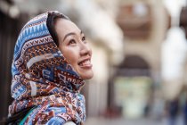 Jovem mulher étnica no lenço de cabeça sorrindo e olhando para longe, enquanto está contra o fundo urbano borrado na cidade de Jeddah, na Arábia Saudita — Fotografia de Stock