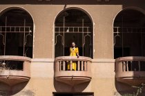 Angolo basso di giovane donna etnica in abito giallo in piedi su un piccolo balcone ad arco di edificio in pietra invecchiata nella città di Jeddah in Arabia Saudita — Foto stock