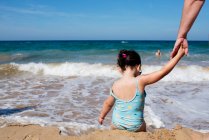 Вид ззаду маленька анонімна дівчинка в купальнику тримає руку батька врожаю, сидячи на мокрій піщаному пляжі і насолоджуючись теплою водою на фоні величного моря — стокове фото
