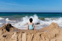 Вид ззаду на невпізнавану маленьку дівчинку в купальнику, що сидить на піщаному пляжі проти морських хвиль і насолоджується літнім відпочинком у сонячний день — стокове фото
