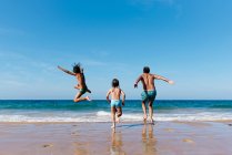 Visão traseira do homem irreconhecível com filhos pequenos correndo e pulando na água do mar enquanto se divertem juntos durante as férias de verão na praia arenosa — Fotografia de Stock